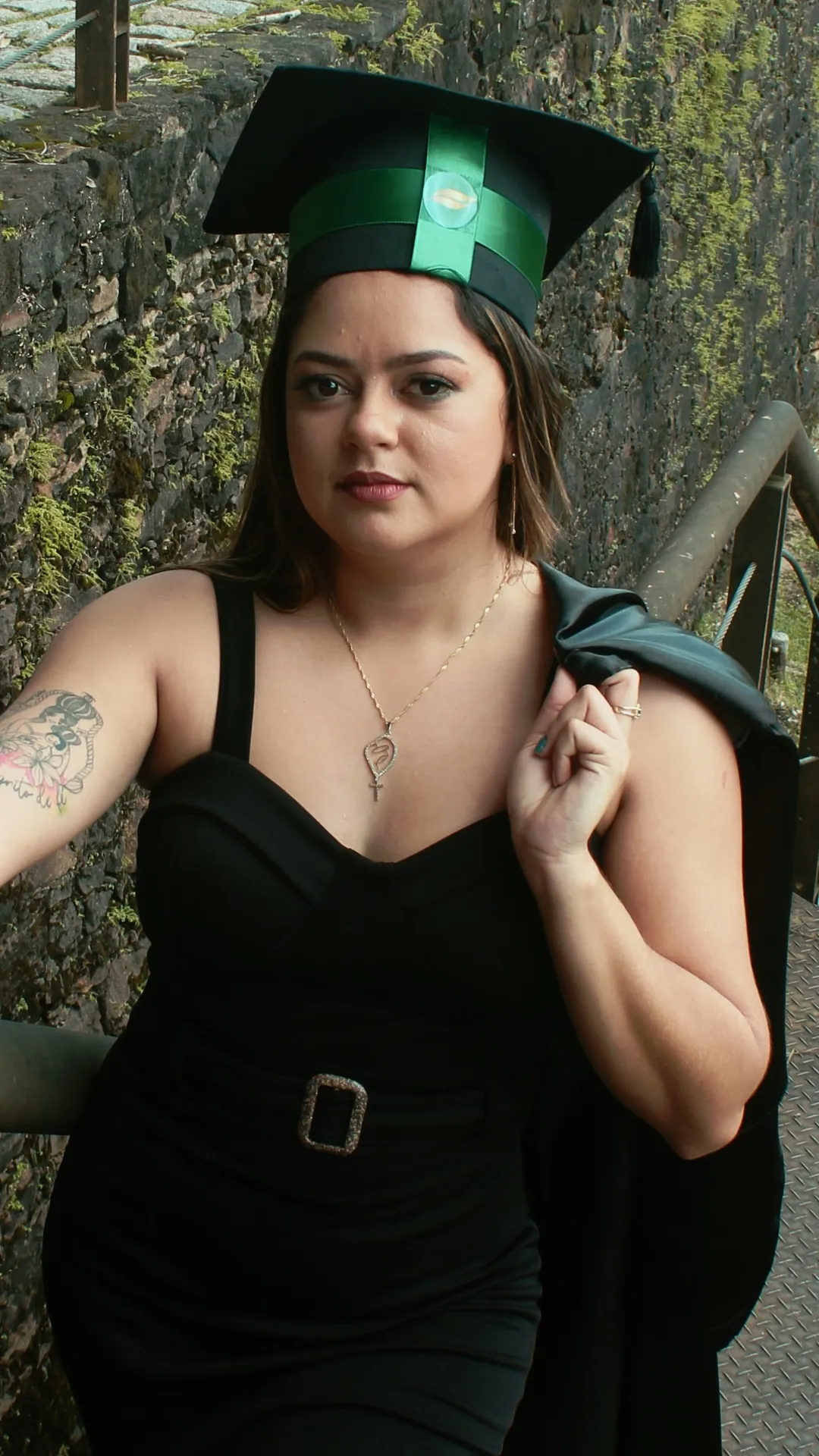 Retrato de formanda de vestido preto com a beca no ombro, olhando para a câmera
