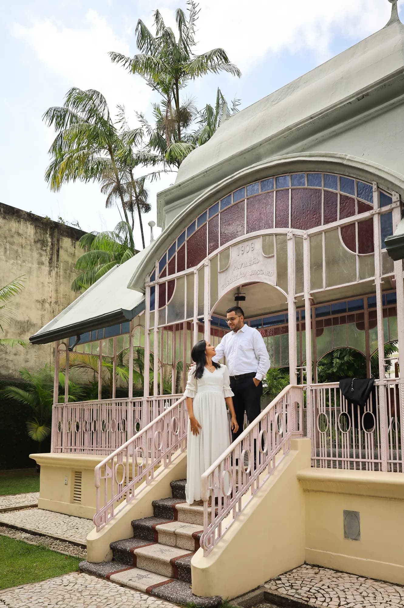 Casal na escadaria de um pavilhão cercado de palmeiras