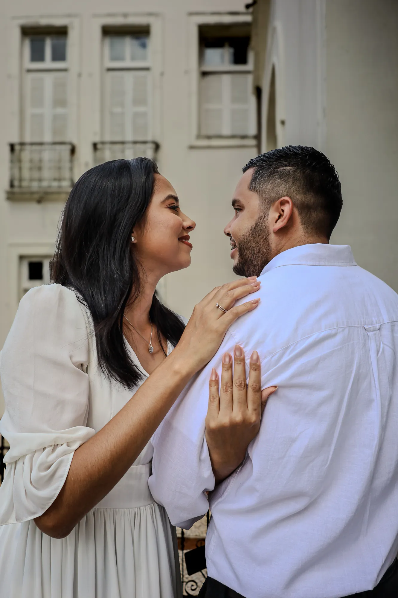 Casal sorrindo e se olhando diante das janelas de um casarão branco