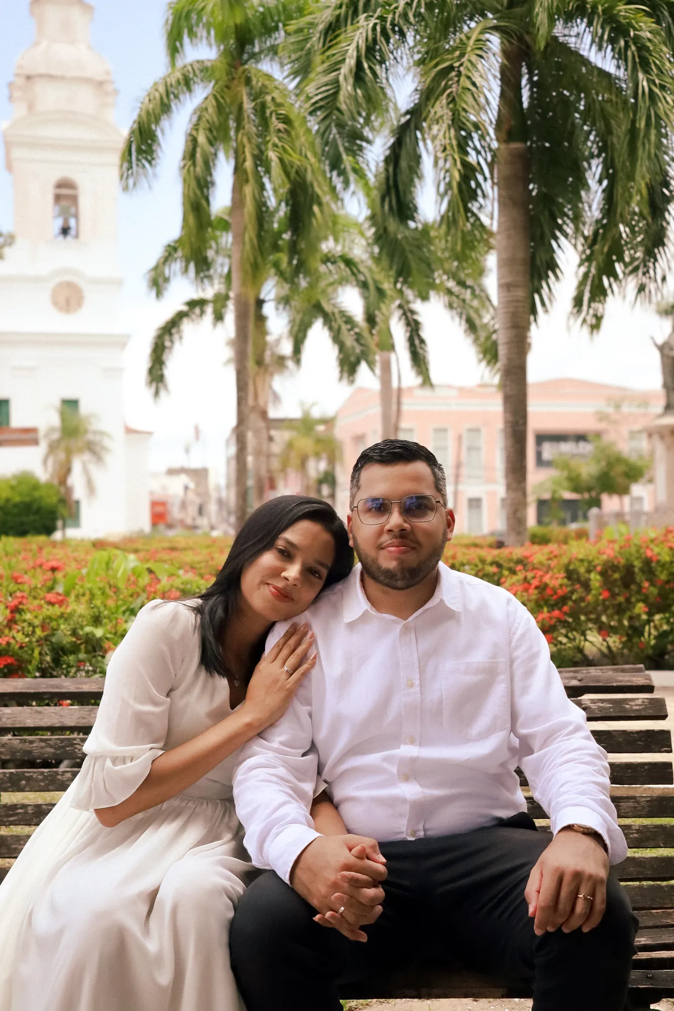 Casal sentado lado a lado em banco de praça, com palmeiras ao fundo