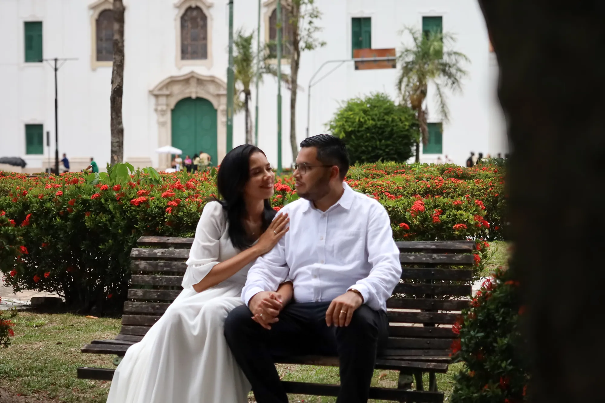 Ensaio pré-wedding em Belém: casal sentado em banco de jardim, cercado de flores vermelhas